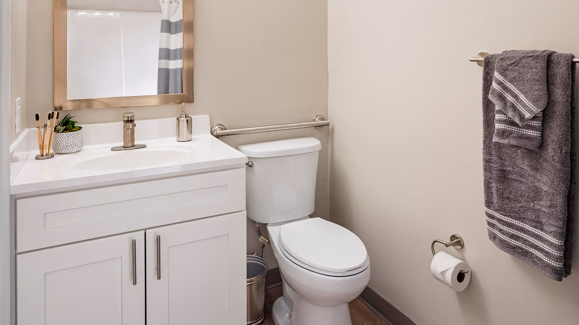 A clean bathroom with a white vanity and sink, a large mirror with a gold frame, a white toilet with a metal grab bar behind it, a metal trash can beside the toilet, and a towel rack on the wall holding two gray towels. A toilet paper holder with a roll of toilet paper is mounted on the wall.