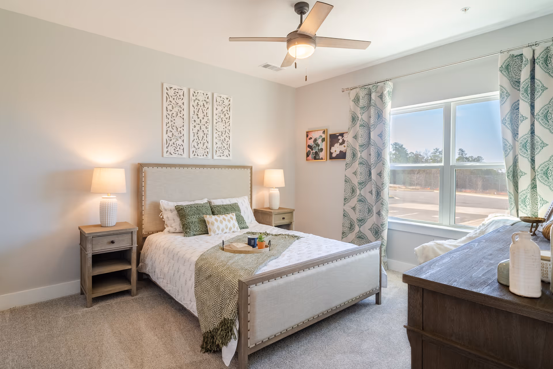 A bright and cozy bedroom with a beige upholstered bed adorned with green and white pillows and a green throw blanket. There are two wooden nightstands with white lamps on either side of the bed. The room has light-colored walls with decorative wall art above the bed and two framed pictures on the adjacent wall. A large window with patterned curtains lets in natural light, and a ceiling fan with a light fixture is mounted on the ceiling. A wooden dresser with decorative items is positioned near the window.