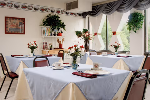 Bright dining room with tables covered in light tablecloths, floral centerpieces and teacups near large windows.