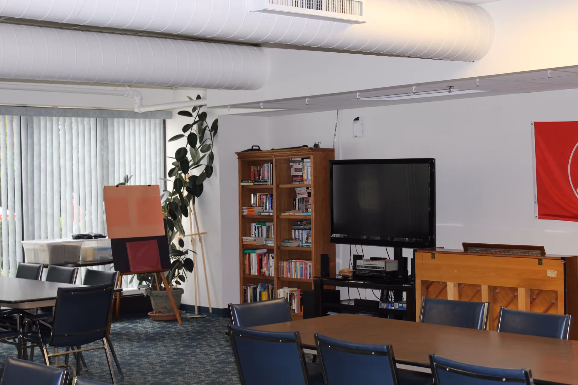Community common room with tables and chairs, a television, bookshelf, piano and an easel by vertical blinds.