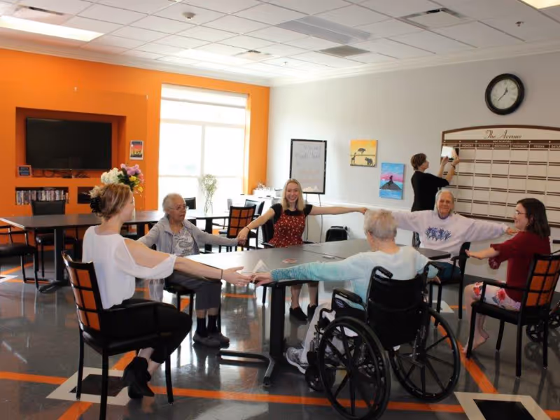 Residents and staff seated around tables in a bright activity room holding hands and doing seated arm-stretch exercises.