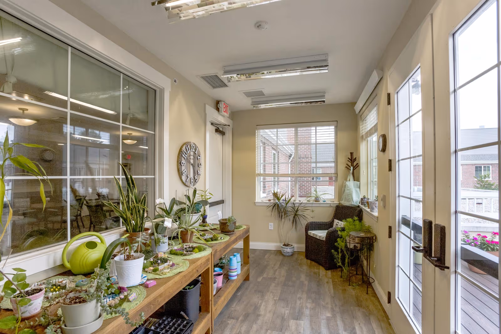A bright indoor sunroom or enclosed porch area with a wooden table holding various potted plants and gardening supplies. There is a green watering can on the table. A wicker chair with a cushion is placed near the window, and a small side table with a plant is next to it. Large windows and glass doors allow natural light to fill the space, and a wall clock is mounted near an exit door.