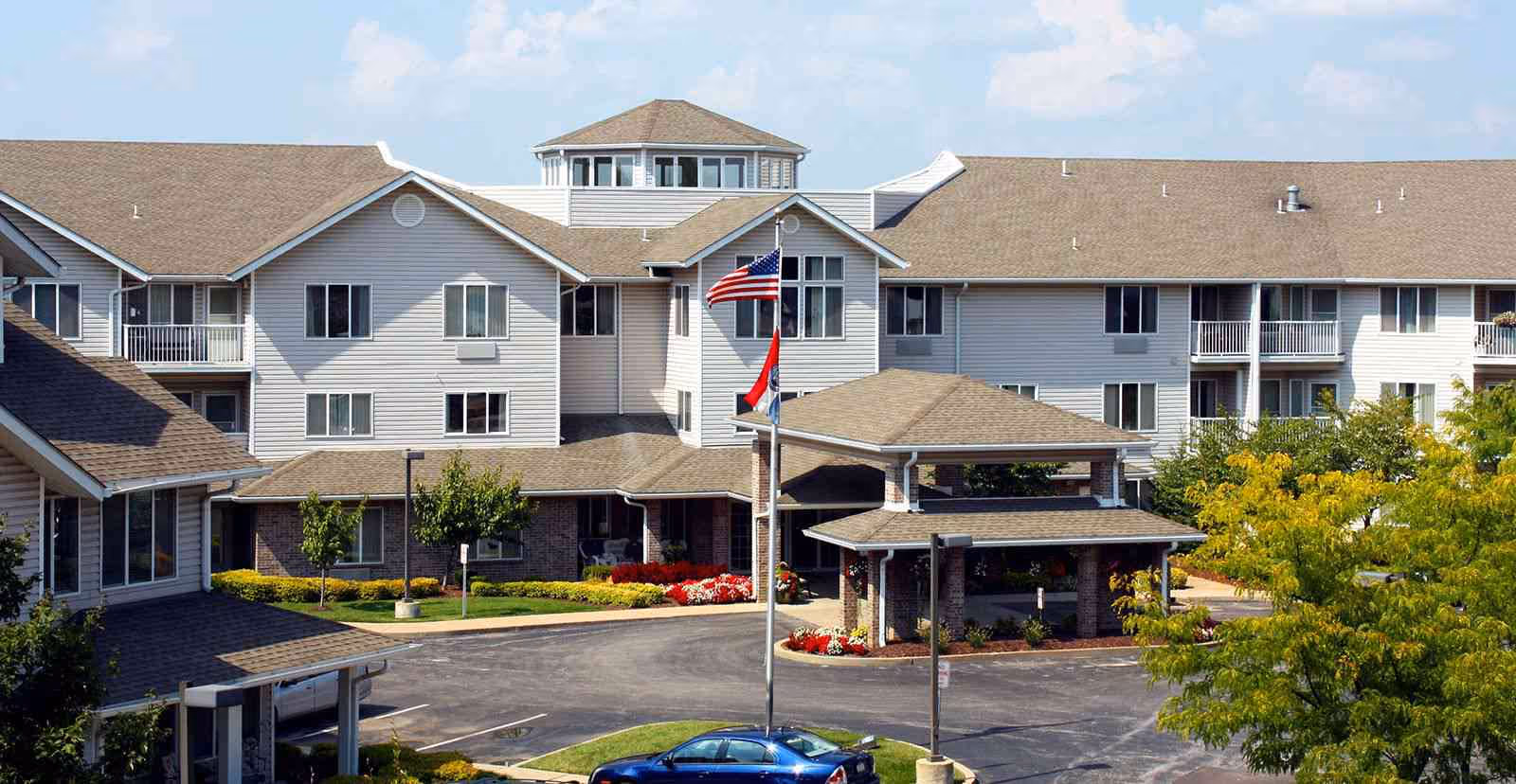 Front exterior of a multi-story senior living facility with a covered porte-cochere, flagpoles, and landscaped entrance.