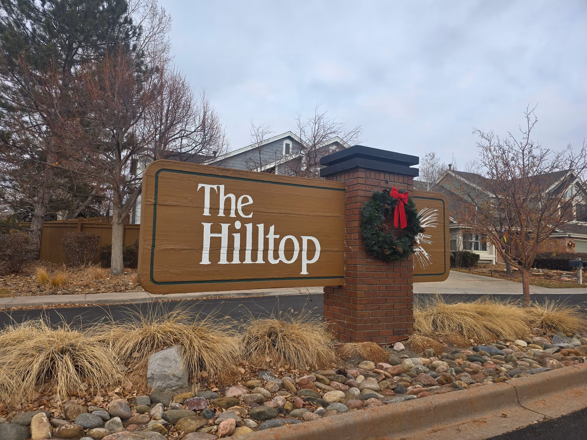 A wooden sign mounted on a brick pillar with a holiday wreath and red bow, displaying the text 'The Hilltop' in white letters. The sign is situated on a landscaped area with rocks and dry grass, with houses and leafless trees in the background under a cloudy sky.