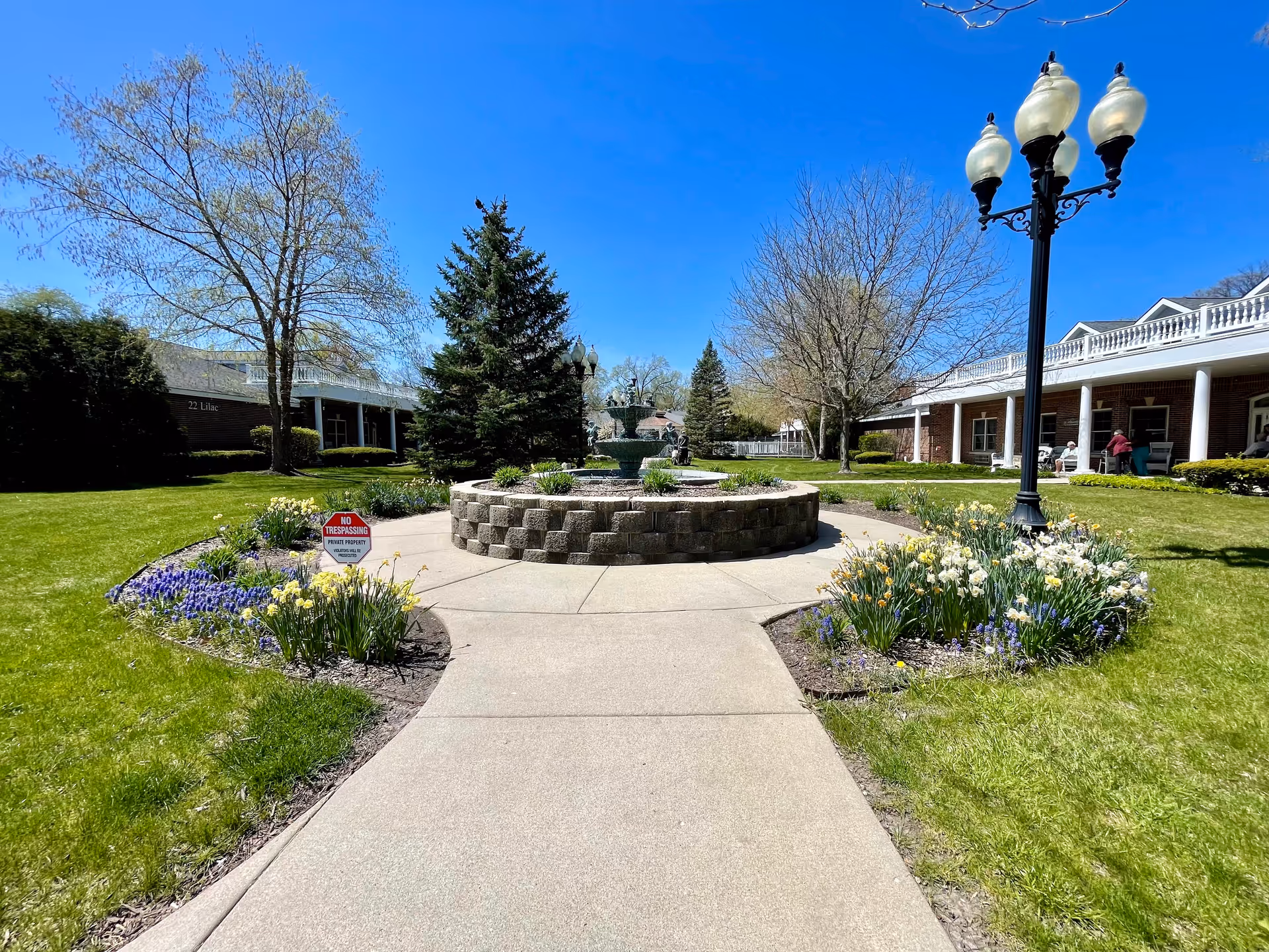 A sunny outdoor courtyard at Cottages of Fox Lake featuring a circular stone fountain surrounded by flower beds with yellow and purple flowers. There are green lawns, leafless and evergreen trees, a black vintage-style street lamp, and brick buildings with white railings and columns in the background under a clear blue sky.