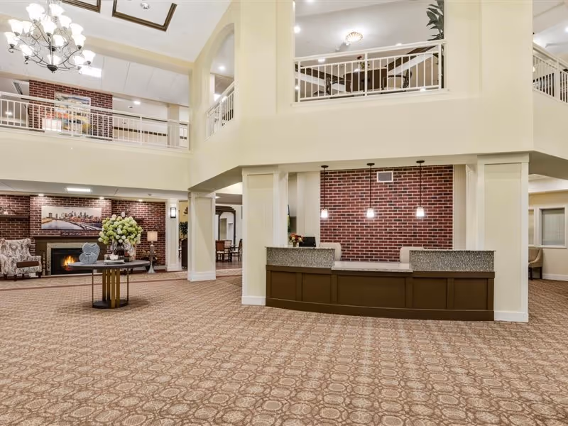Spacious senior living facility lobby with a patterned carpet, a reception desk with three hanging pendant lights, a round table with a flower arrangement, a fireplace with a cityscape painting above it, and seating areas with armchairs. The lobby has a high ceiling with a chandelier and a second-floor balcony overlooking the area.