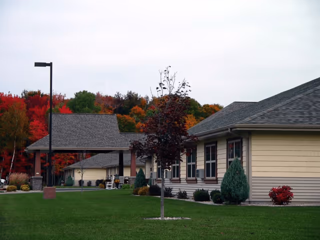 Single-story beige senior living building with a covered entrance, manicured lawn, and colorful autumn trees in the background.