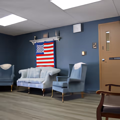 A sitting area with two blue armchairs and a blue sofa against a blue wall. Above the sofa is a shelf with small decorative items and an American flag hanging on the wall. There is a wooden door with a small window on the right side of the image. The floor is light-colored wood, and the ceiling has white fluorescent lights.