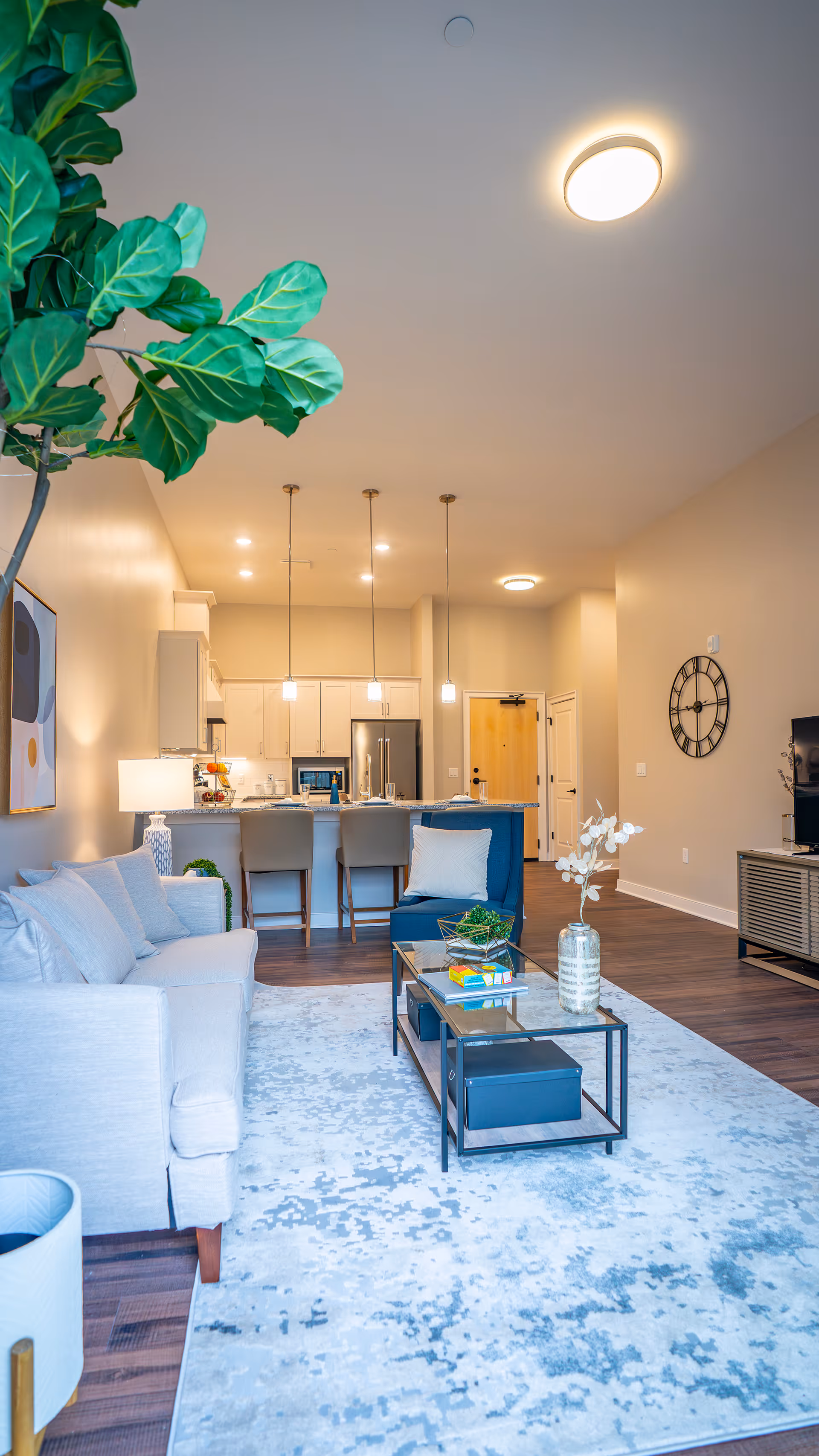 Bright open-plan living room and kitchen with a sofa, coffee table on a patterned rug, bar stools at an island, and modern pendant lighting.