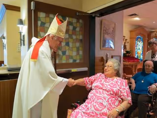 A clergyman dressed in white religious robes and a mitre is shaking hands with an elderly woman in a pink floral dress seated in a wheelchair inside a nursing home. Another elderly woman in a blue top is seated nearby, and a caregiver stands behind them. The setting appears to be a common area with stained glass and framed artwork on the walls.