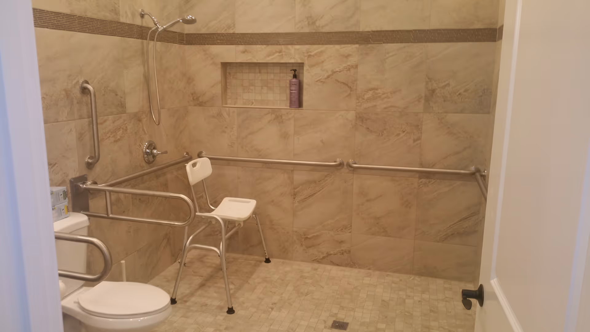 Accessible bathroom with beige tiled walls and floor, equipped with a white shower chair, multiple stainless steel grab bars around the toilet and shower area, a wall-mounted showerhead, and a recessed shelf holding a bottle of soap or shampoo.