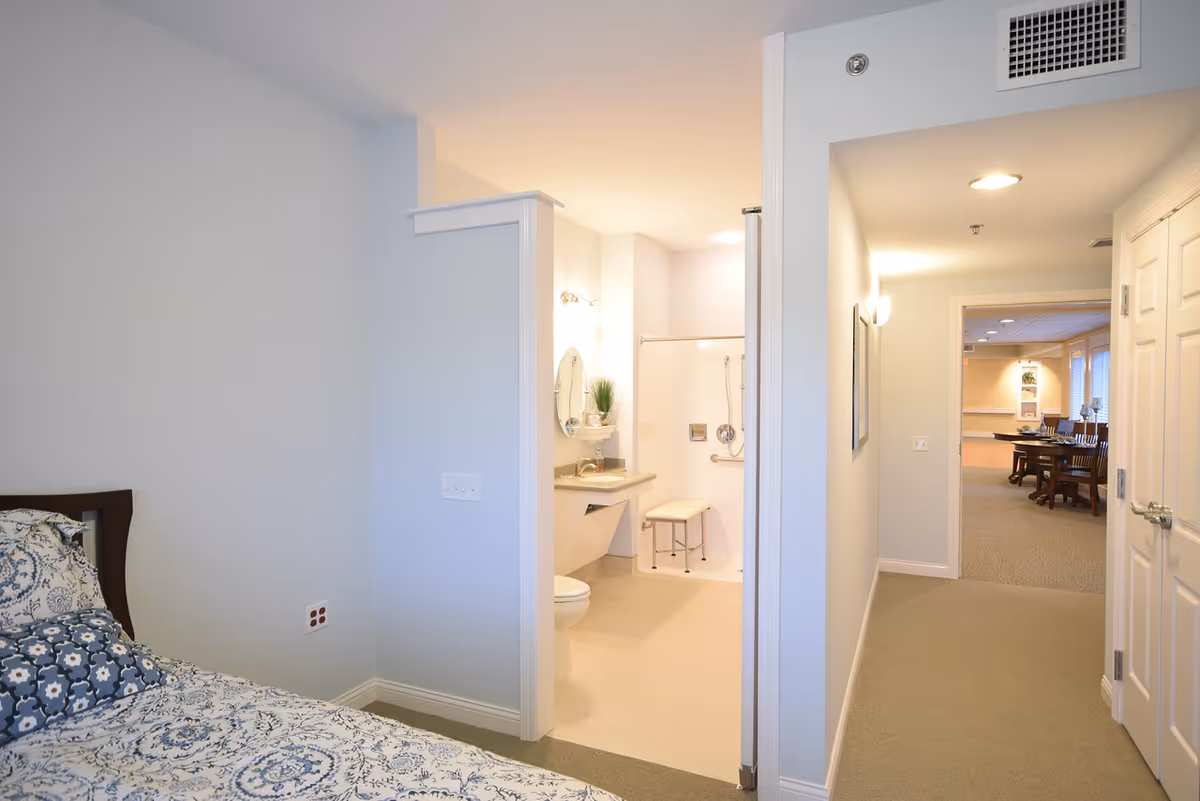 View of a senior living facility interior showing a bedroom with a bed covered in patterned bedding on the left, an adjacent bathroom with a toilet, sink, mirror, and shower with a bench, and a hallway leading to a dining area with tables and chairs in the background.