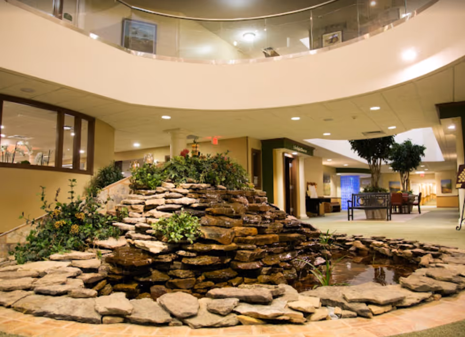 Indoor water feature with stacked stones and plants in the center of a spacious retirement community lobby area with seating and large potted trees, under a circular balcony with glass railing.