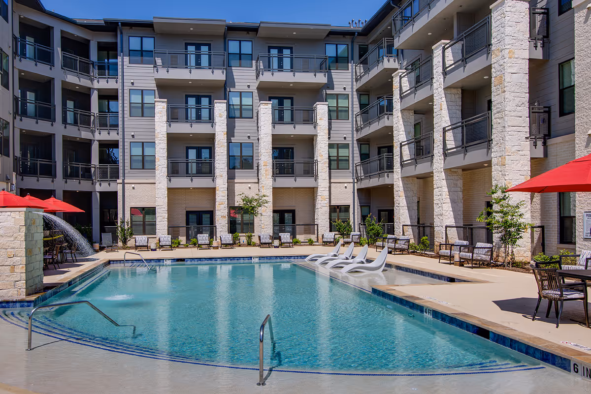 Outdoor swimming pool area at Fountainwood at Lake Houston with lounge chairs partially submerged in the water, red umbrellas providing shade, and a multi-story residential building with balconies surrounding the pool.
