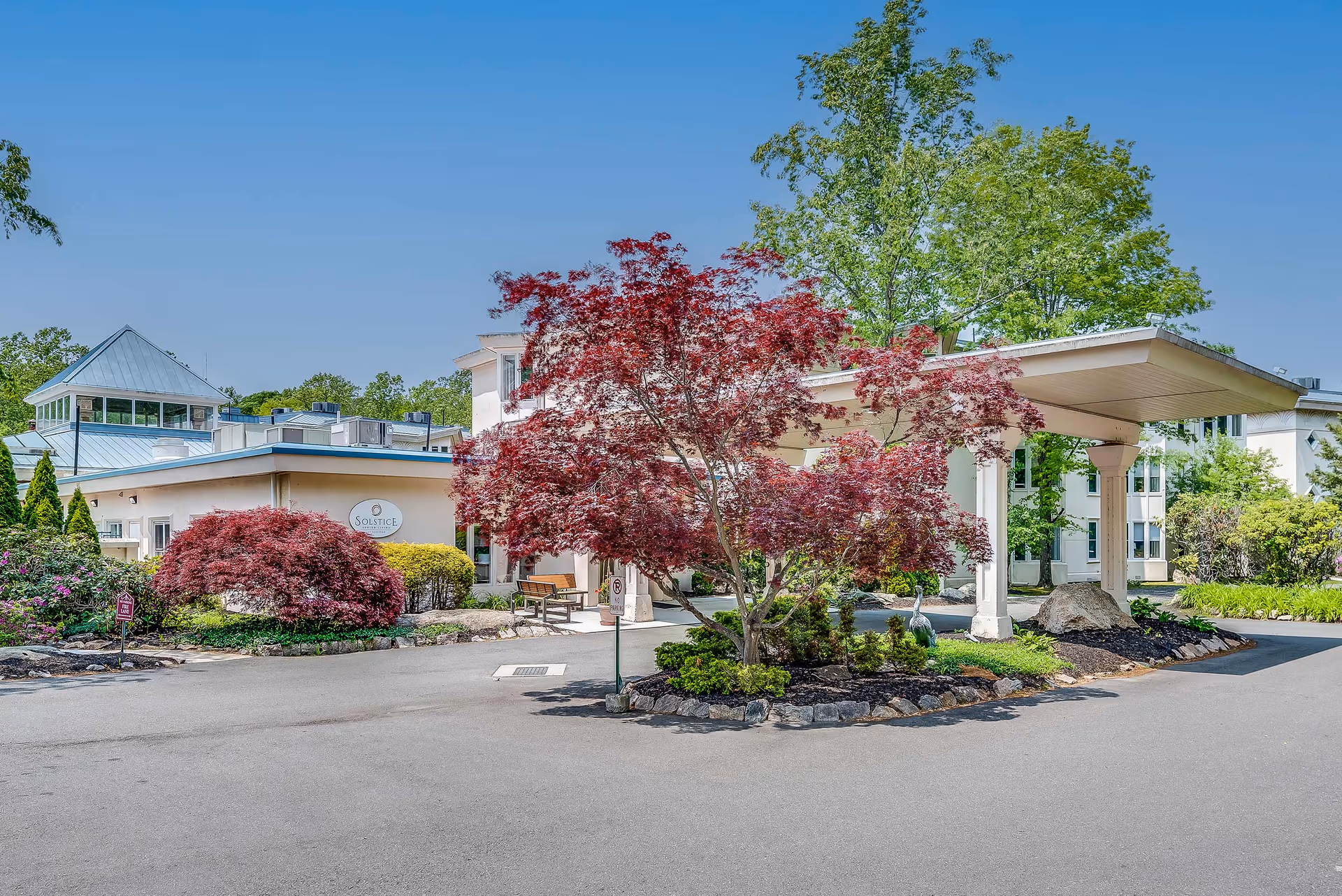Exterior view of a retirement residence building with a covered entrance driveway. There are well-maintained landscaping elements including red and green trees and shrubs around the driveway. The building has light-colored walls and multiple windows, with a clear blue sky in the background.