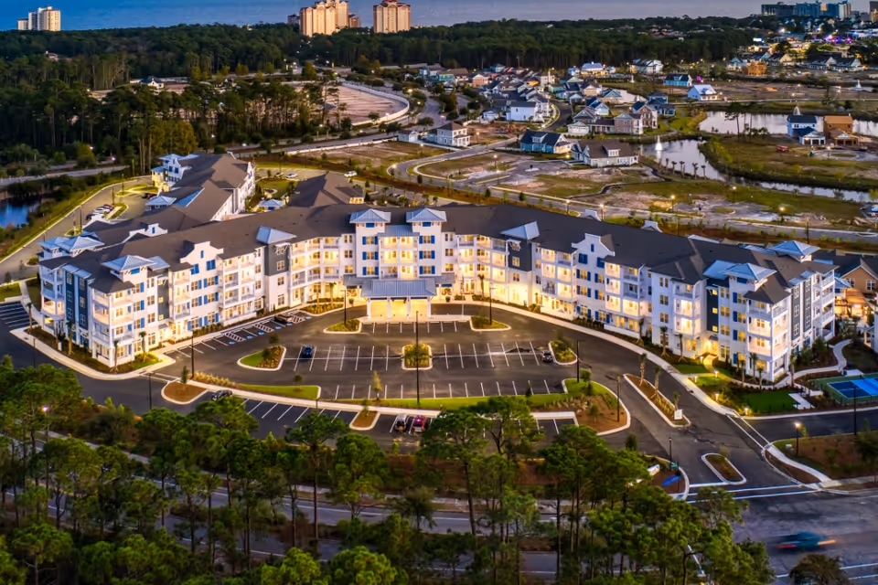 Aerial evening view of the illuminated, multi-story Portside at Grande Dunes building with a curved facade, parking lot, and surrounding landscaping.