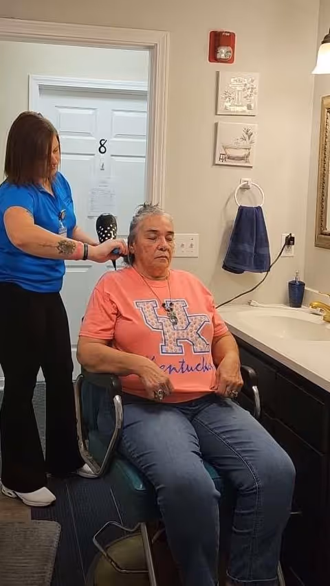 A woman wearing a pink University of Kentucky t-shirt sits in a chair while another woman in a blue shirt styles her hair with a hairdryer in a room with a sink, towel, and wall decorations.