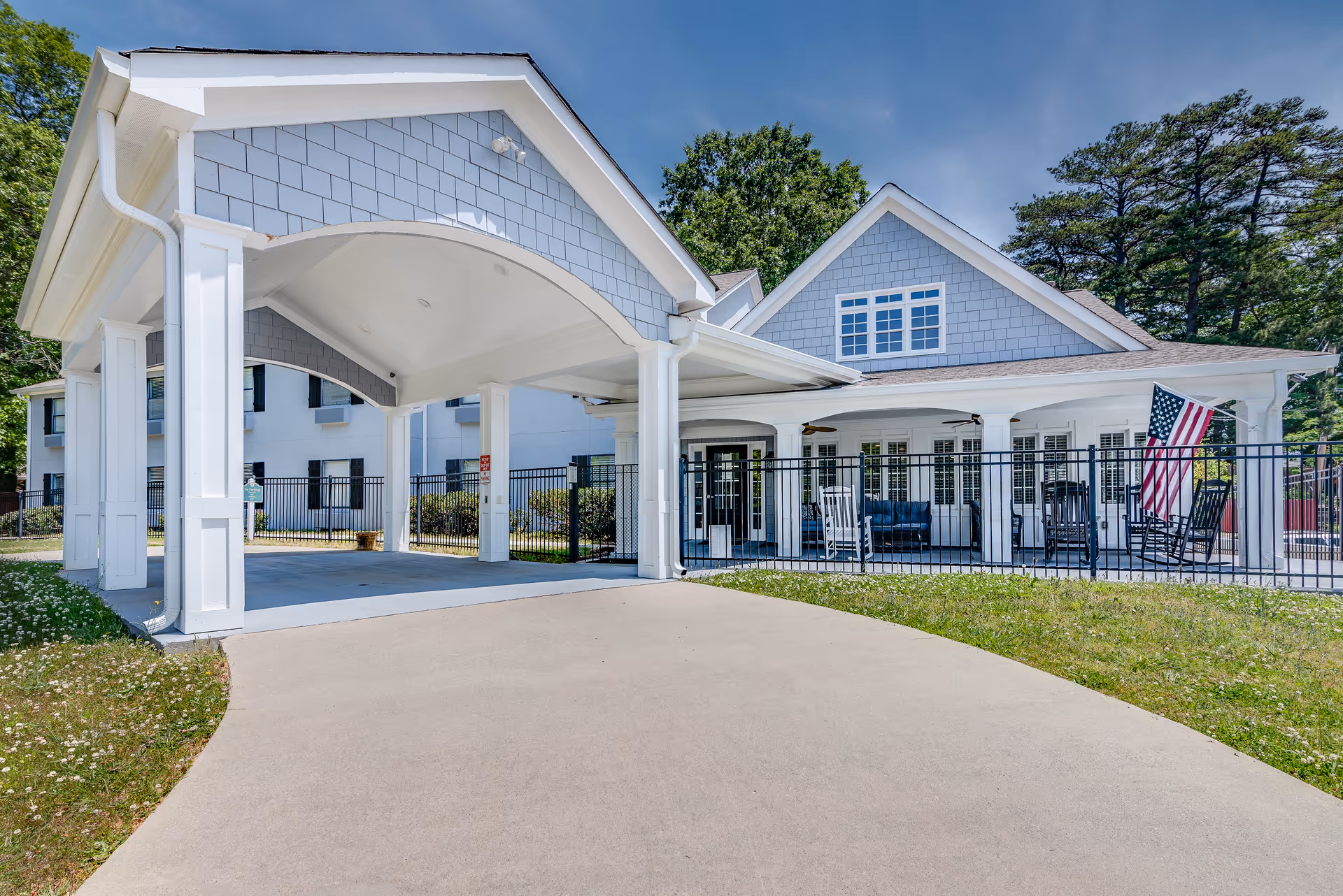 Exterior view of a senior living facility named Enrich @ 519 - Senior Living, showing a covered driveway entrance with white pillars and a blue-gray building with white trim. There is a fenced patio area with rocking chairs and an American flag, surrounded by green grass and trees under a clear blue sky.