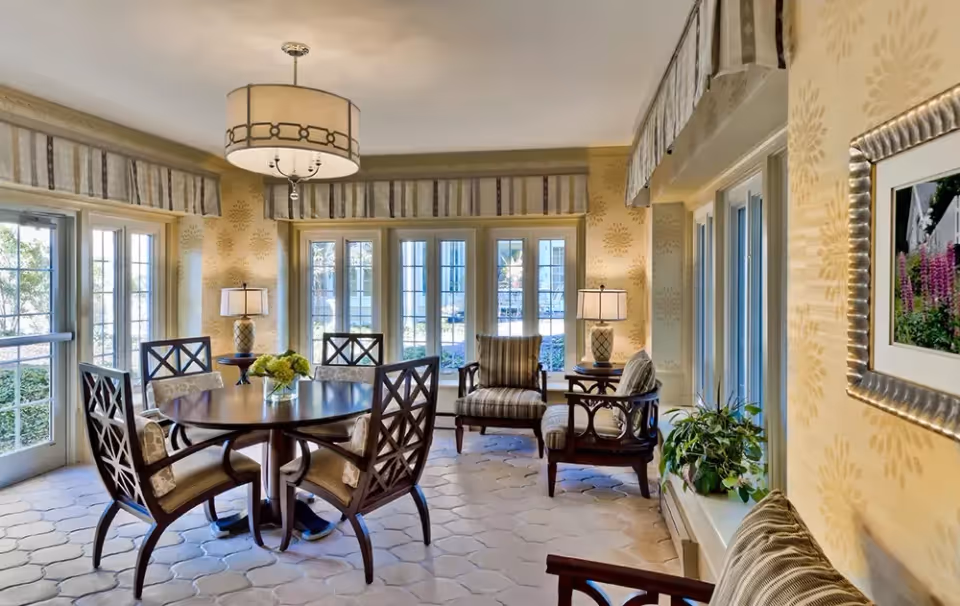 A bright and cozy interior room with large windows and glass doors letting in natural light. The room features a round wooden dining table with four cushioned chairs, two armchairs with striped cushions, side tables with lamps, a hanging ceiling light fixture, and a framed floral picture on the wall. There is also a small green plant on a windowsill.