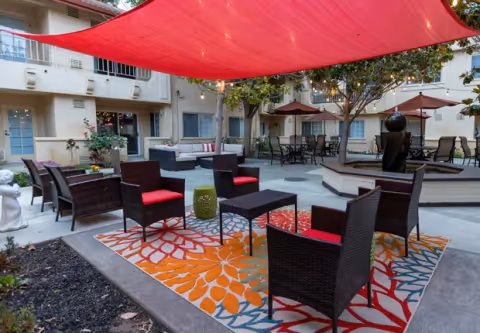Outdoor courtyard area of a senior living facility with wicker chairs featuring red cushions arranged around a small black table on a colorful floral-patterned rug. There are additional seating areas with tables and umbrellas, string lights overhead, and a water fountain surrounded by trees. The building with balconies and windows surrounds the courtyard.