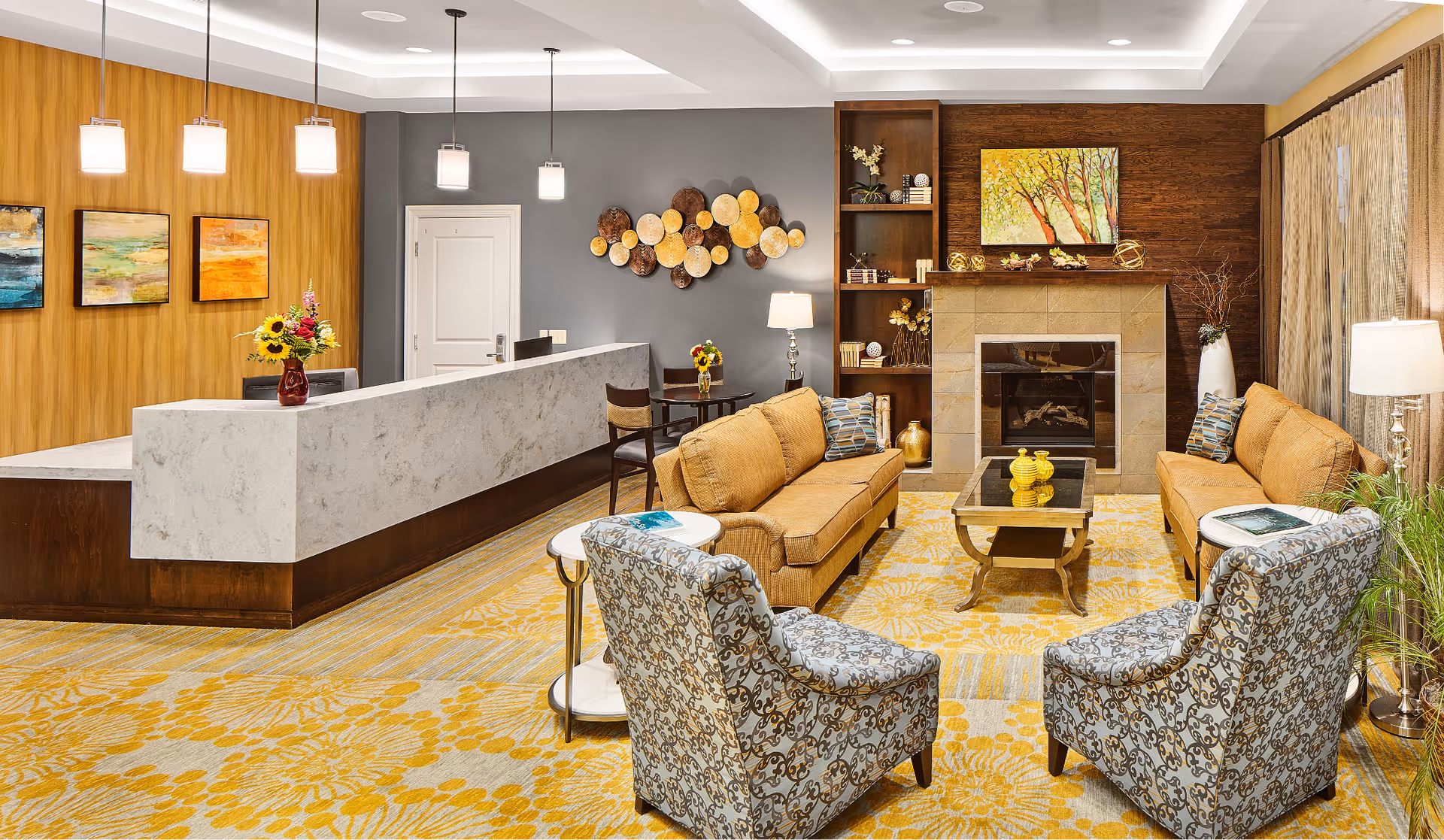 A cozy and well-lit living room area in a senior living facility featuring two mustard-colored sofas facing each other with patterned armchairs, a glass coffee table, a fireplace with a painting above it, and a reception desk with pendant lights and decorative wall art in the background.