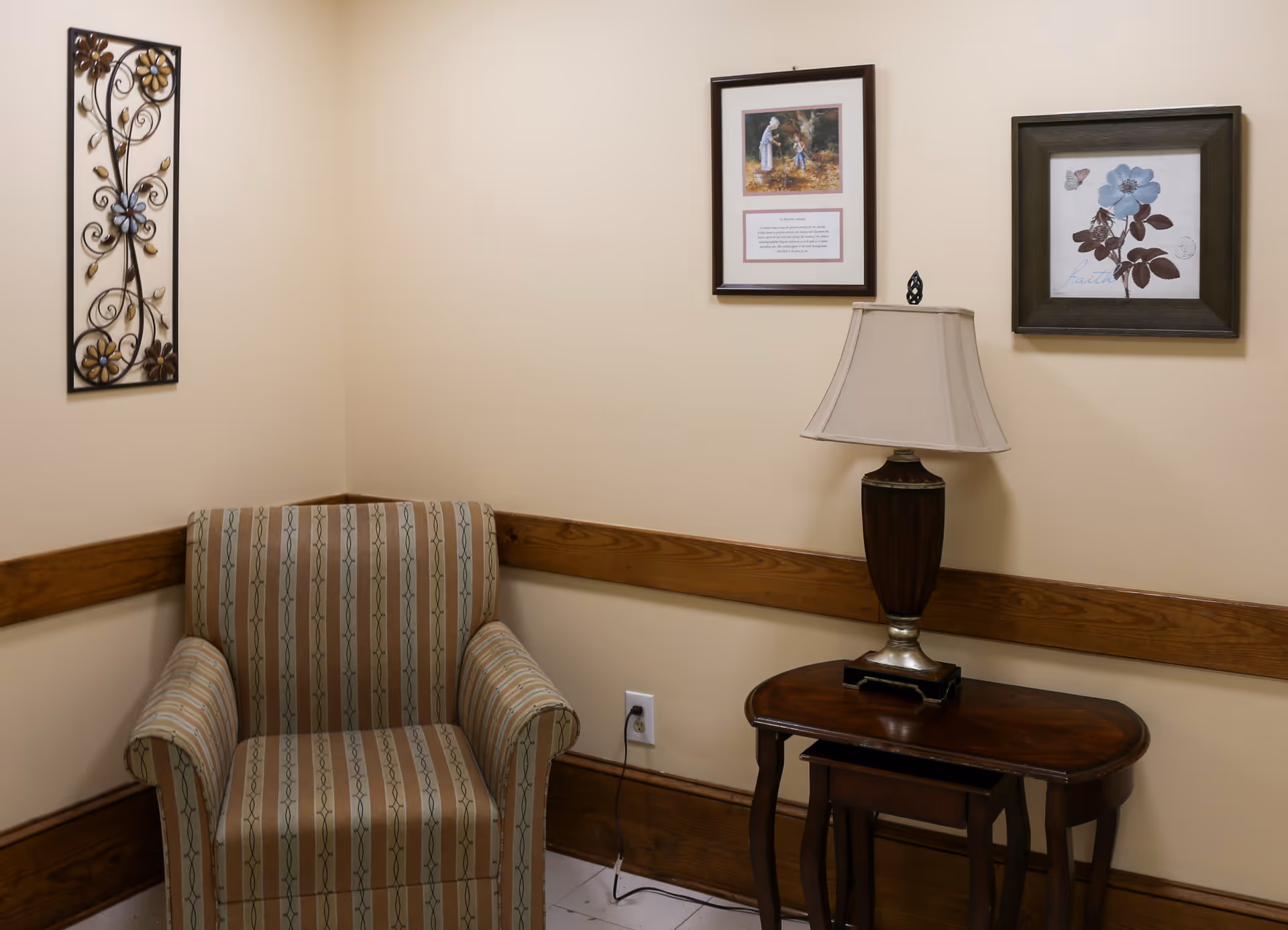 A cozy corner in a room with a striped armchair, a wooden side table with a lamp, and three framed wall decorations including a floral metal art piece and two pictures with flowers and a person.