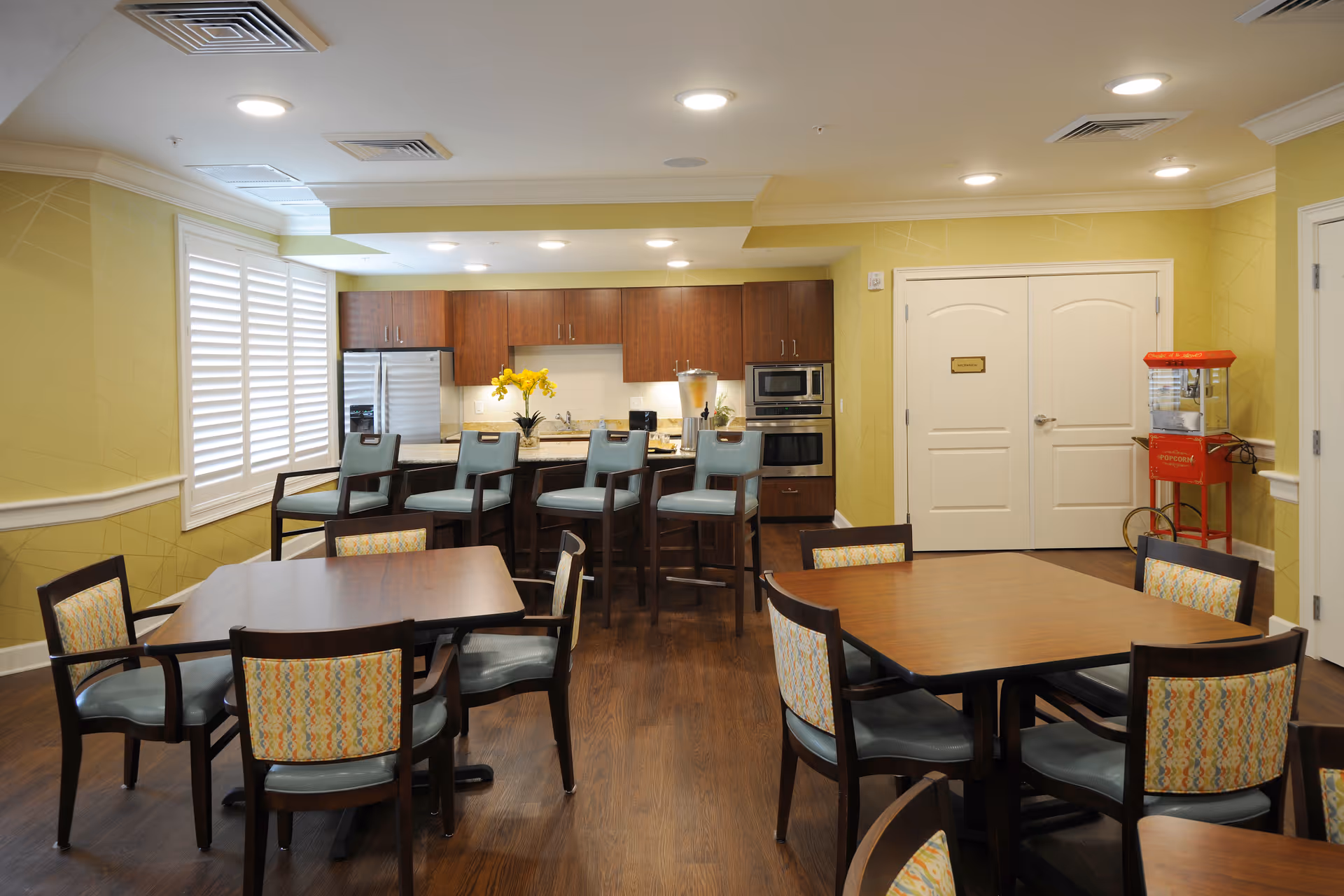 A bright dining area in a senior living facility featuring wooden tables with patterned cushioned chairs. In the background, there is a kitchen area with wooden cabinets, a stainless steel refrigerator, microwave, and oven. Four high chairs are placed at the kitchen counter. A red popcorn machine is positioned near double white doors on the right side. The room has yellow walls and wood flooring.