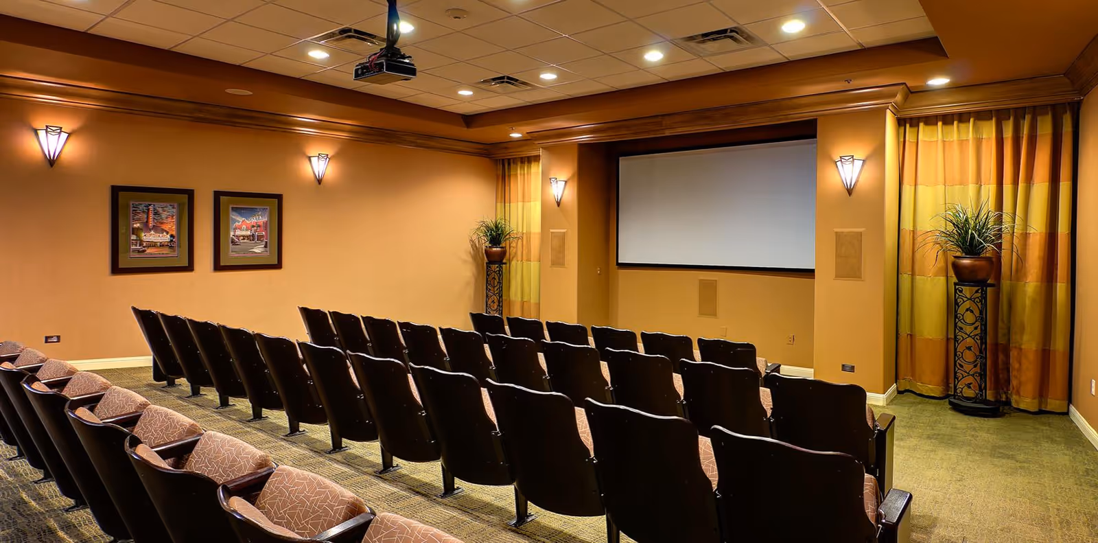 Small screening room with rows of theater chairs facing a projection screen, warm lighting, framed art, and potted plants.
