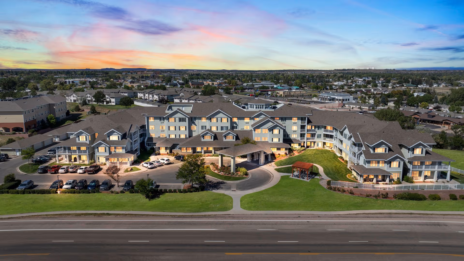 Aerial view of Whispering Chase | Sky Active Living senior living facility at sunset, showing a large multi-wing building with multiple windows lit from inside, surrounded by green lawns, a parking lot with cars, and a small gazebo on the grass. The background includes a suburban neighborhood and a colorful sky with clouds.