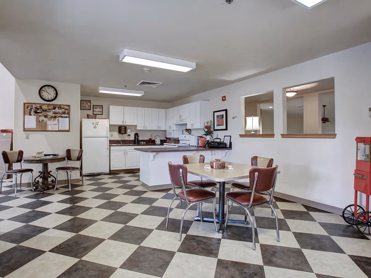 A senior living facility common area featuring a kitchen with white cabinets, a refrigerator, and a stove. In front of the kitchen is a dining area with a square table and four brown chairs. The floor has a black and white checkered pattern. There is a small round table with two chairs on the left side and a red popcorn machine on the right. The walls are white with two open windows looking into another room.