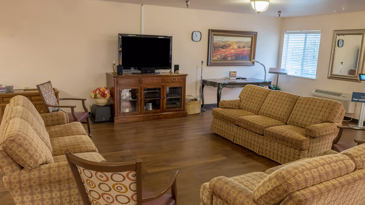 A senior living community common area with patterned upholstered sofas and chairs arranged around a wooden TV cabinet holding a flat-screen television. The room has wooden flooring, a wall clock, a framed landscape painting, a window with blinds, a mirror, and a small table with a lamp and some items on it.