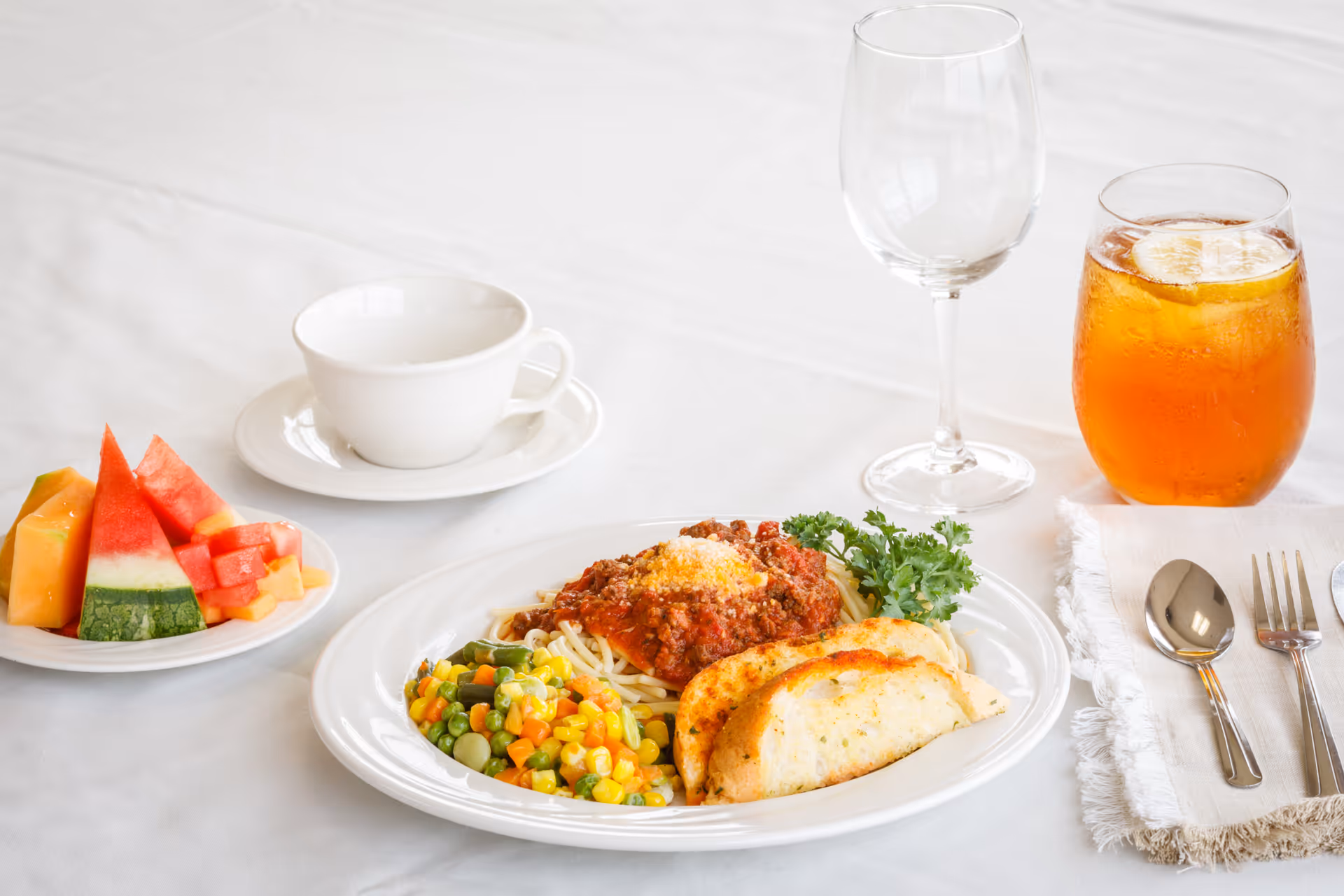 Table setting on a white tablecloth with a plated meal of spaghetti and meat sauce, mixed vegetables and garlic bread, a small fruit plate, a cup, a wine glass, iced tea, and utensils.