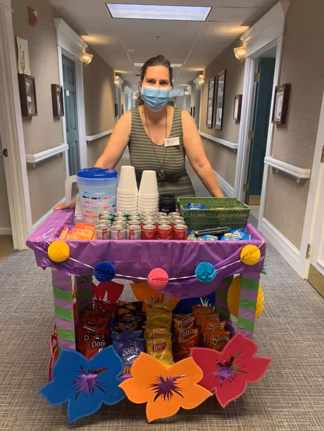 A woman wearing a face mask and a striped sleeveless top stands behind a decorated snack cart in a hallway. The cart is covered with a purple cloth and adorned with colorful paper flowers and round decorations. On the cart are various snacks including bags of chips, cans of soda, disposable cups, and a large water pitcher. The hallway has beige walls, framed pictures, and several doors.