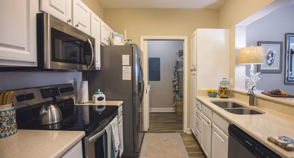 A modern kitchen with white cabinets, a stainless steel microwave, stove, and refrigerator. There is a double sink with a lamp and decorative items on the counter. The kitchen opens into another room with a laundry area visible in the background.