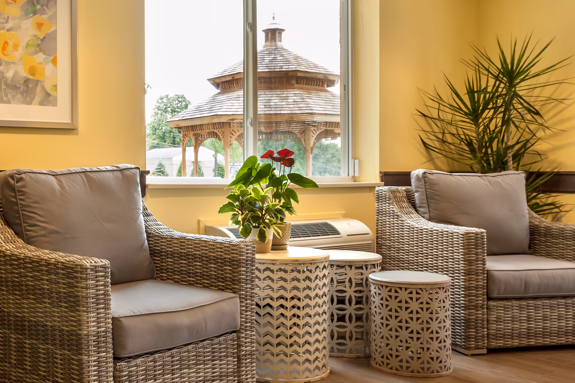 Two wicker armchairs with cushions flank decorative nesting tables and a potted plant in front of a window showing a gazebo outside.