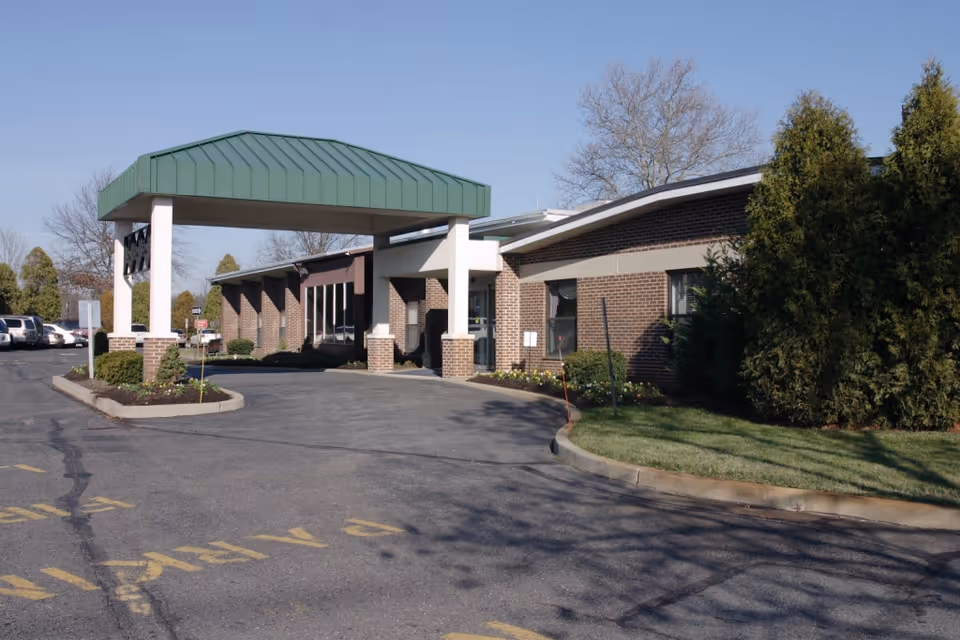Front exterior of a single-story brick senior care building with a green metal covered entrance, driveway, parked cars, and landscaped beds.