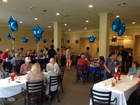 A large room with multiple tables covered with white and blue tablecloths, decorated with blue star-shaped balloons. Several elderly people are seated and standing, engaging in conversation and activities, suggesting a social gathering or event in a communal dining or activity area.