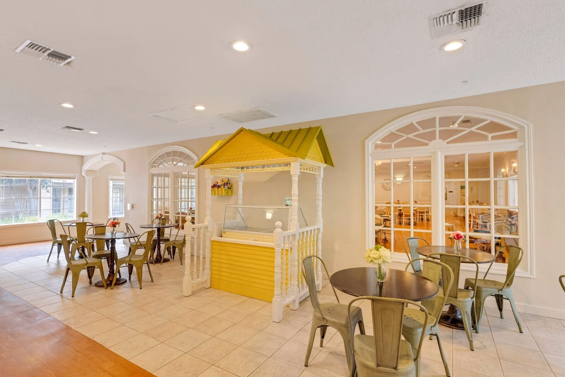 A bright and clean dining area in a senior living facility with several round tables and metal chairs. There is a small yellow kiosk or stand with a green roof in the center of the room. Large windows and glass doors provide a view into an adjacent room with more tables and chairs. The floor is tiled, and the walls are painted light beige.