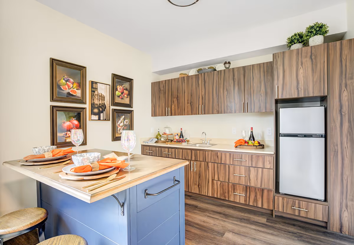 A modern kitchen with wood grain cabinets, a small refrigerator, and a sink. There is a blue kitchen island with two place settings including plates, bowls, orange napkins, and wine glasses. Four framed pictures of fruits and wine bottles hang on the wall.