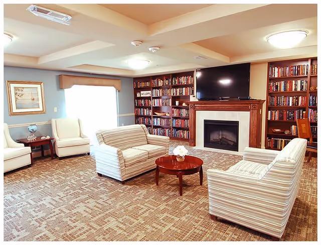 A cozy living room with striped armchairs and a loveseat arranged around a round wooden coffee table with a small flower arrangement. The room features a fireplace with a flat-screen TV mounted above it, flanked by built-in bookshelves filled with books. There are two white armchairs near a window with a framed picture on the wall and a small side table with a globe and decorative items.