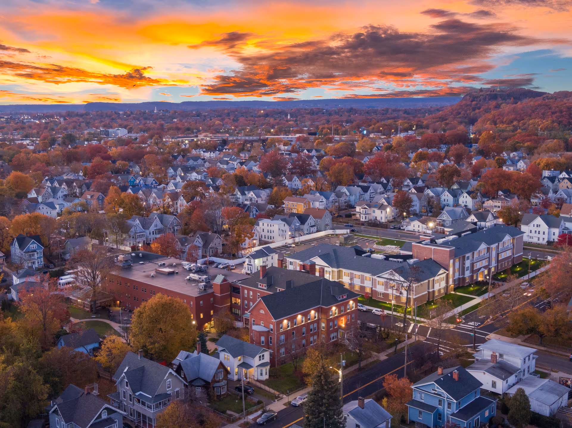 Aerial view of a residential neighborhood at sunset with a large senior living facility named Mary Wade prominently visible in the foreground surrounded by houses and autumn trees under a colorful sky.