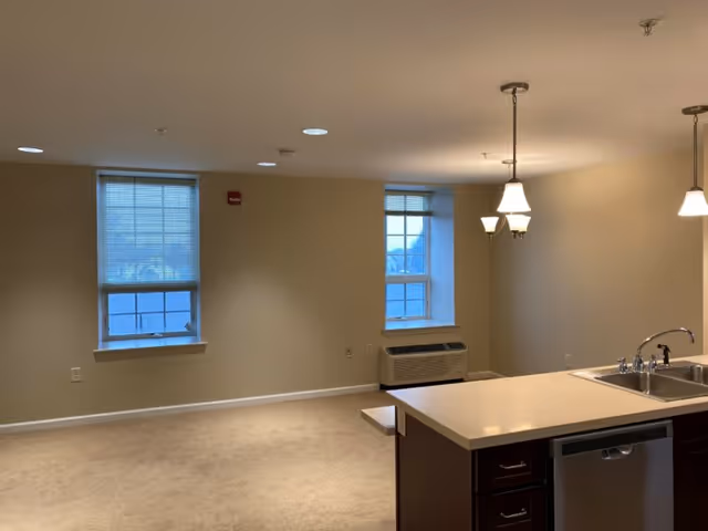 Interior view of a senior living apartment with beige walls and carpet. Two windows with blinds allow natural light into the room. A kitchen island with a sink and dishwasher is visible in the foreground, with hanging light fixtures above. The room appears clean and empty, ready for furnishing.