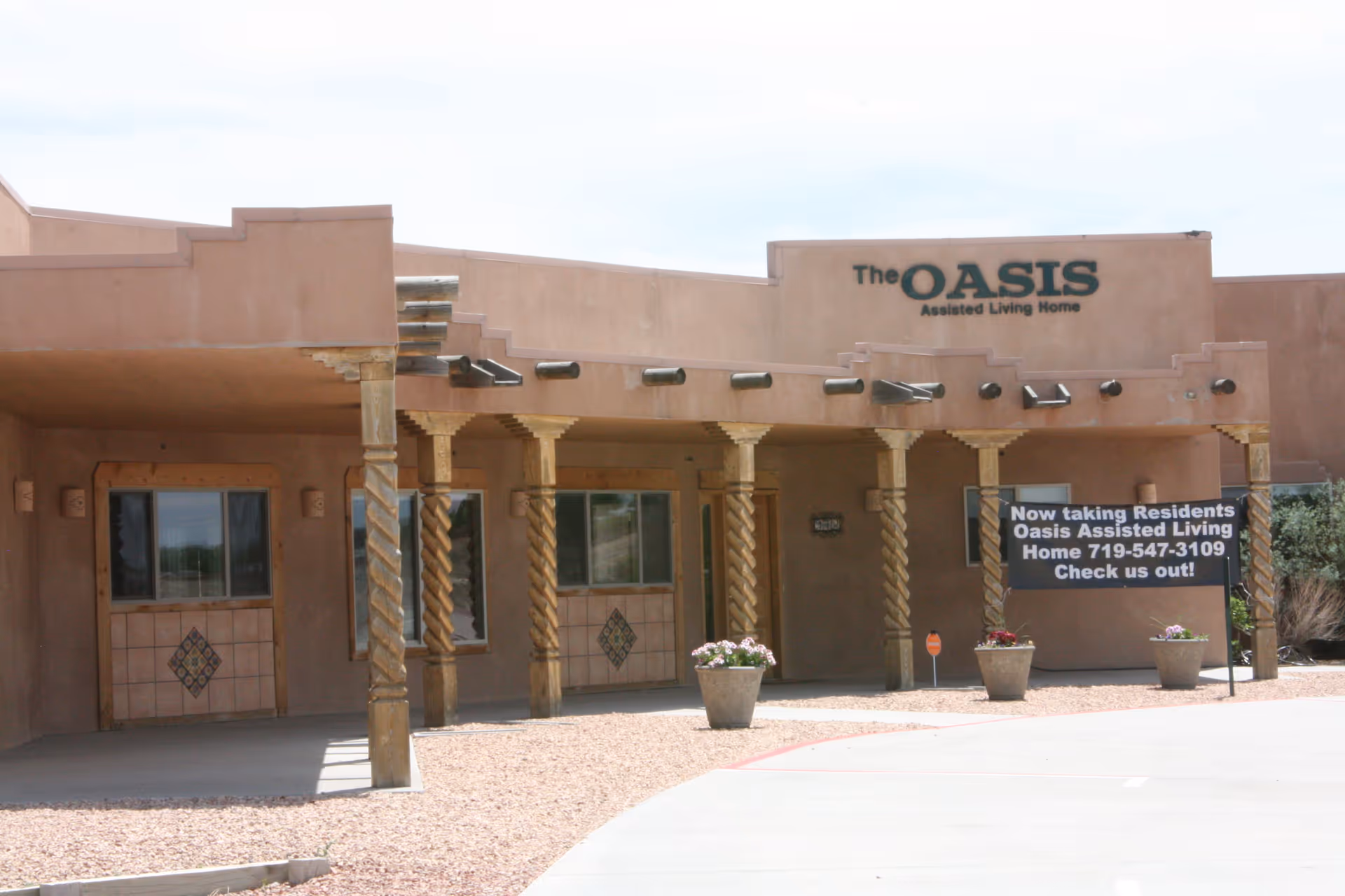 Exterior view of The Oasis Assisted Living Home building with southwestern architectural style, featuring wooden twisted columns, tiled wall decorations, potted plants, and a sign indicating they are now taking residents with a contact phone number.