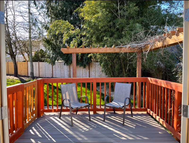 A wooden deck with red railing and two gray outdoor chairs. The deck overlooks a green grassy yard with trees and a wooden fence in the background. A wooden pergola structure is partially visible above the deck.