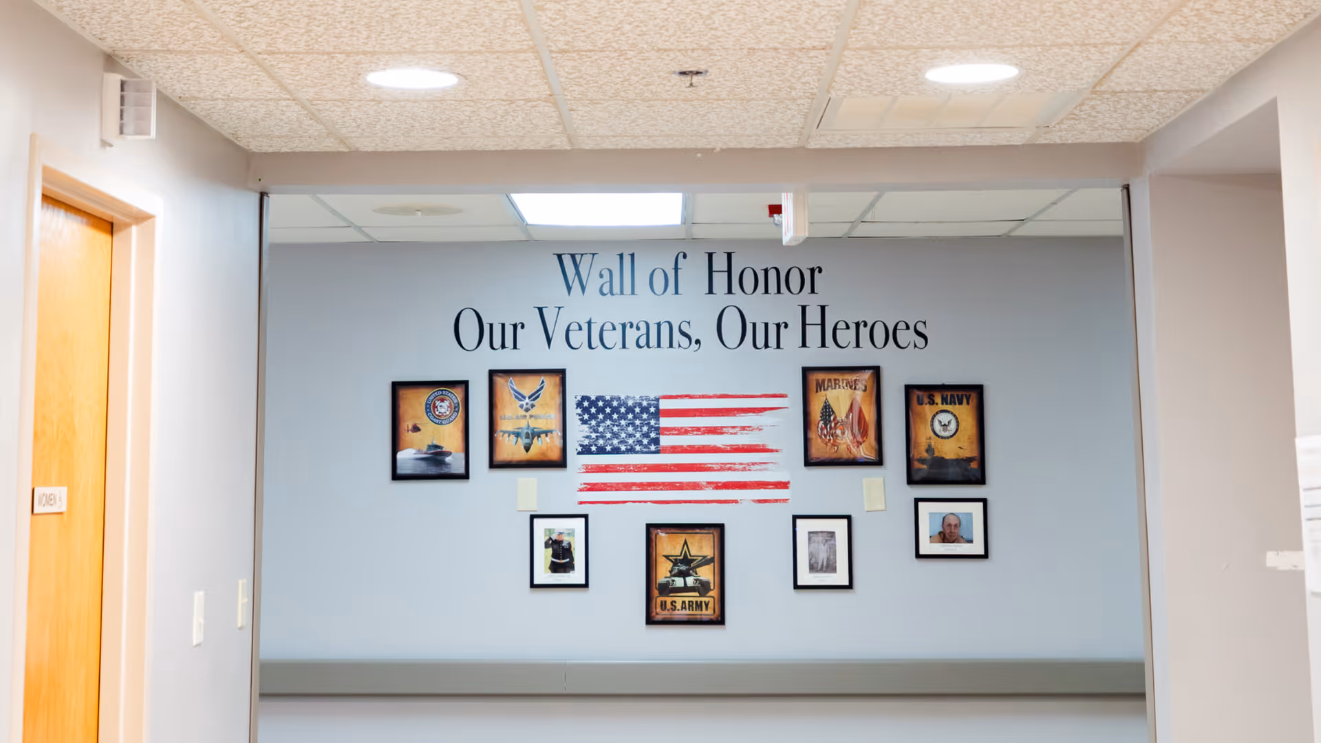 A hallway wall display titled 'Wall of Honor Our Veterans, Our Heroes' featuring framed pictures and emblems representing different branches of the U.S. military, including the Air Force, Marines, Navy, and Army, with an American flag graphic in the center.