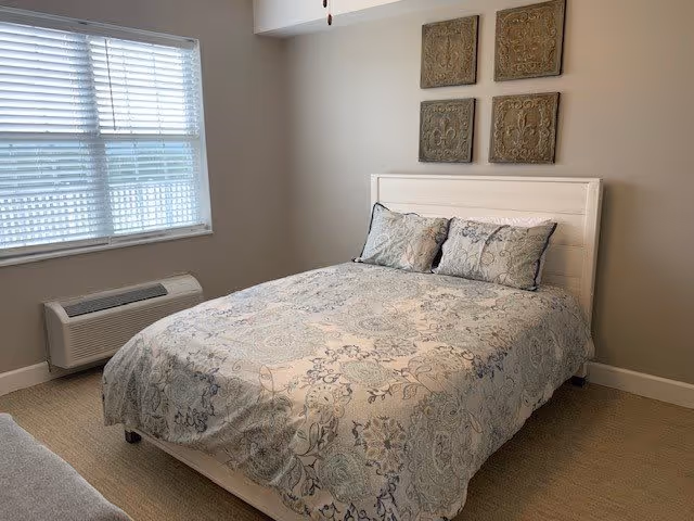 A bedroom with a bed covered in a patterned comforter and two matching pillows. The bed has a white headboard. Above the bed, there are four decorative square wall hangings. To the left, there is a window with white blinds and a heating/cooling unit below it. The walls are painted light beige and the floor is carpeted.