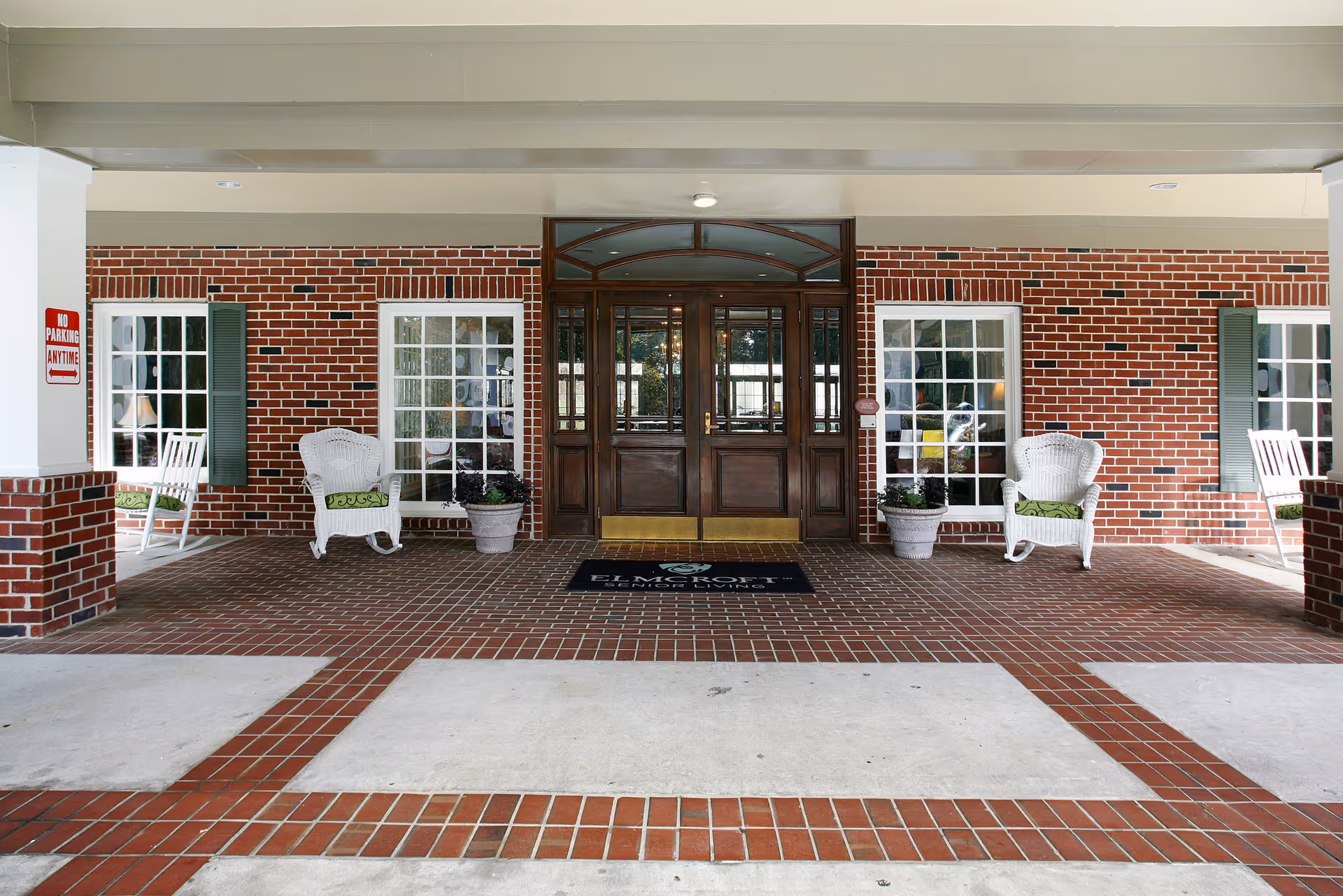Covered entrance of a building with a brick facade, featuring double wooden doors with glass panels, two white wicker chairs with green cushions, potted plants, and large windows with green shutters on either side. The floor is tiled with red bricks and concrete sections, and a 'No Parking Anytime' sign is visible on the left pillar.