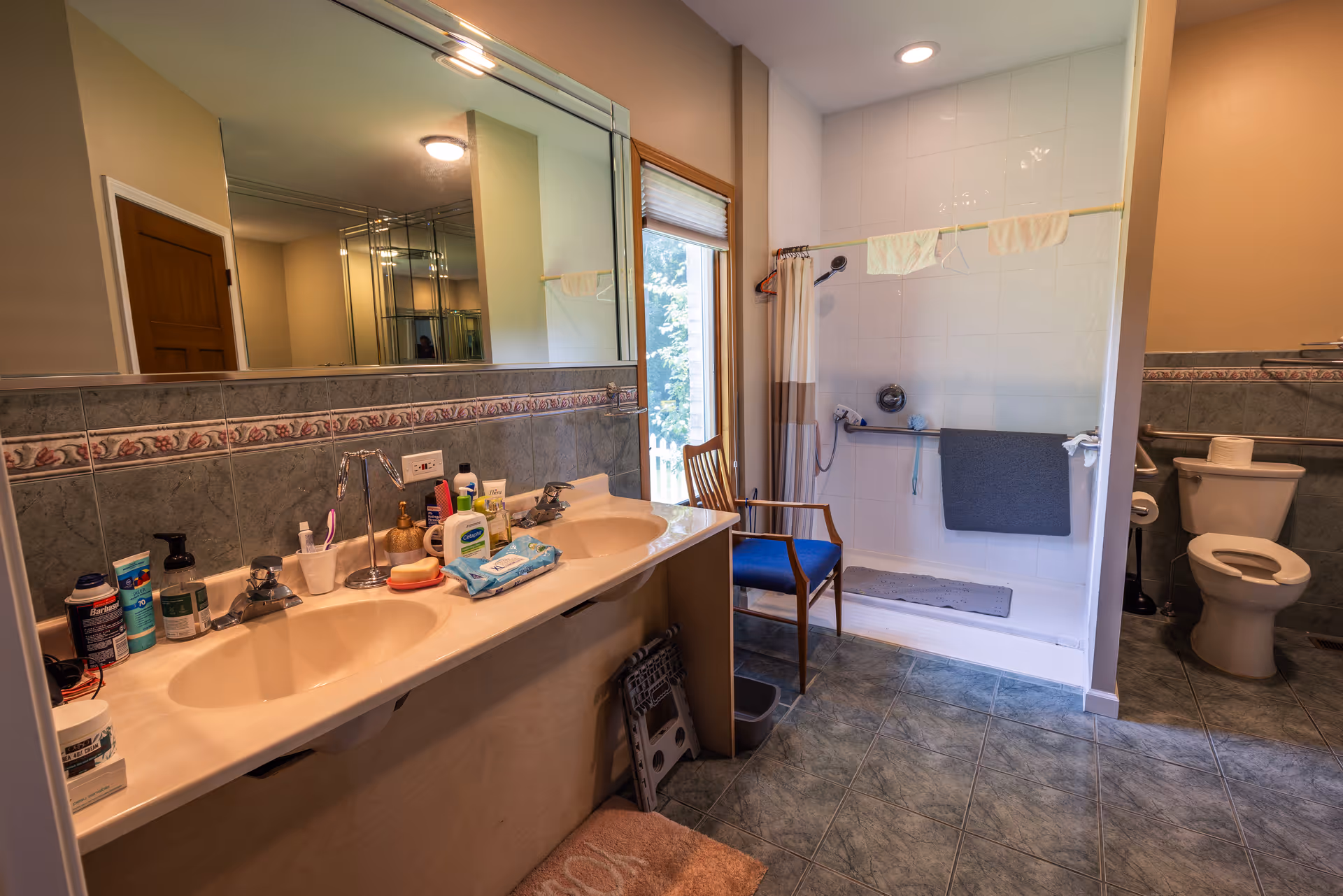 Bathroom with a double sink vanity cluttered with toiletries, a large mirror above the sinks, a walk-in shower with a chair inside, and a toilet in the corner. The floor is tiled and there is a window next to the shower letting in natural light.