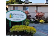 Exterior view of a senior living facility building with a sign in front that reads 'Greenridge Senior Care 2150 Pineland Drive'. The building is surrounded by bushes and small trees under a clear sky.