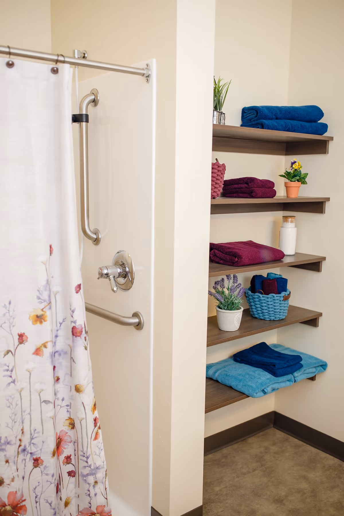 A bathroom shower area with a floral patterned shower curtain partially drawn to the side. The shower has a metal grab bar and a control knob. Adjacent to the shower are wooden shelves mounted on the wall, holding neatly folded towels in blue and purple colors, small potted plants, and a jar.